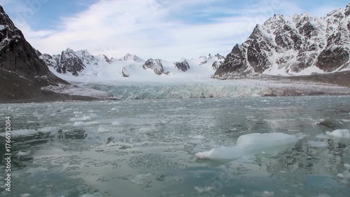 A calm Arctic ocean filled with icebergs. Mountains covered in snow and ice rise dramatically behind a glacier in Svalbard. The glacier is slowly melting into the sea.