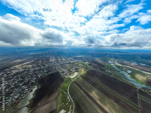 Expansive, high aerial perspective of villages and a distant urban area nestled within a vast, striped plain under a huge sky filled with bright, dramatic clouds