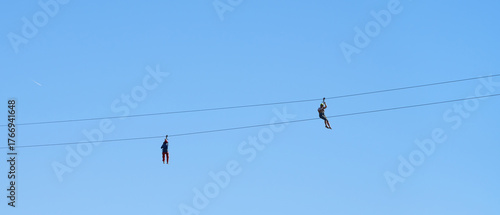Ziplining. Two female figures on a metal cable suspended in the air against a blue sky.