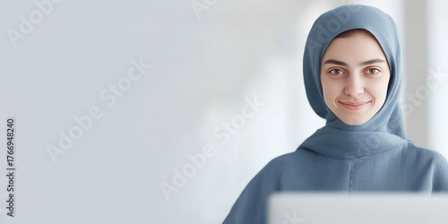 Young woman wearing a blue hijab is smiling while working on a laptop in a bright, modern workspace, showcasing professionalism and confidence in a serene environment