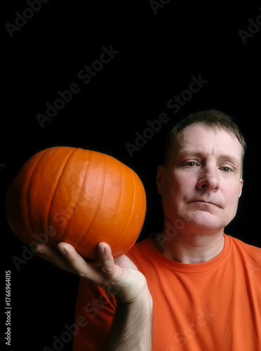 Serious man wearing orange shirt holding a bright orange pumpkin on his palm against a dark black background. Minimalistic Halloween or autumn concept symbolizing the season, harvest, and festive mood