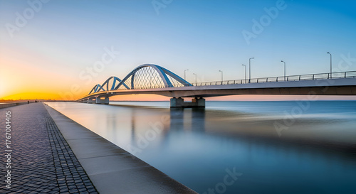 Modern arch bridge spanning a calm river at sunset with a clear blue sky and golden sun on the horizon