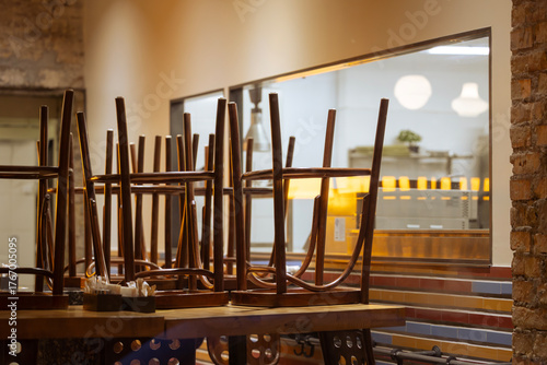 Chairs are stacked upside down on wooden tables in a restaurant after closing time. The kitchen is visible through a large window in the background. This scene captures the quiet end of a business day