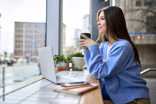 Happy calm lady student enjoying warm hot drink with mug in hands working or studying online. Young smiling woman sitting holding cup drinking tea or coffee relaxing in cafe looking through window