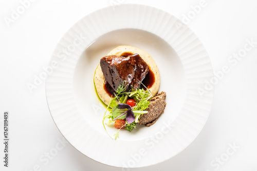 A gourmet dish of braised beef, possibly short ribs, on a bed of creamy purée, served with a small fresh salad and a dark, rich sauce, on a white ridged plate.