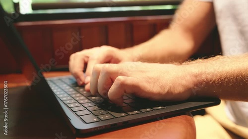 Male hands typing on a keyboard while working on a laptop on a wooden table with warm lights  