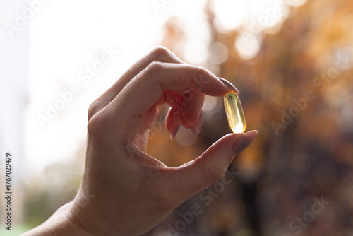 Closeup of a person holding a vitamin D 3 capsule against colourful trees in fall.