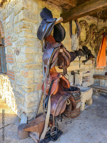 Rustic collection of worn, brown leather saddles and black tack hanging on a wooden rack against a rough, historic stone wall in a stable or rural tack room