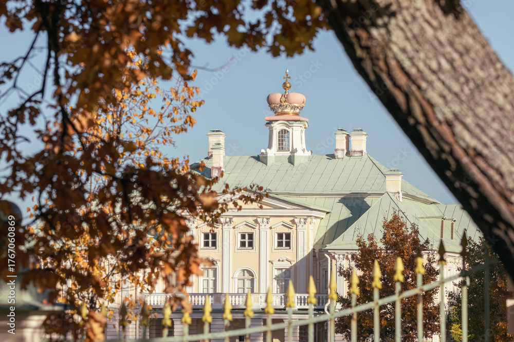 Obraz premium Close view of Grand Menshikov Palace in Oranienbaum framed by autumn leaves and decorative fence under clear blue sky.