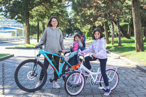 Smiling woman with two children on bicycles in sunny park alley, one child sitting in bike seat. Family enjoying an active outdoor weekend together. Concept of healthy lifestyle, leisure, family time