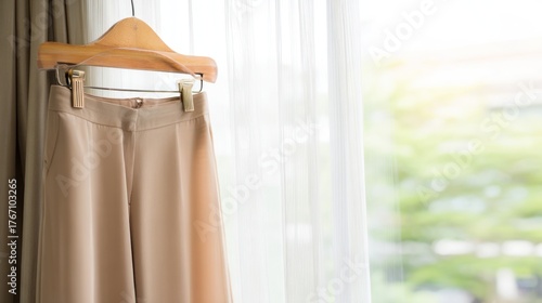 Tan trousers hanging on wooden hanger by window with natural light