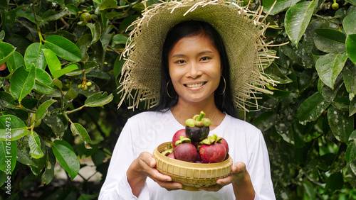 Portrait, smiling southeast asian woman farmer with harvested purple mangosteen tropical fruits on farm or plantation, tree in background, looking at camera, in indonesia, asia