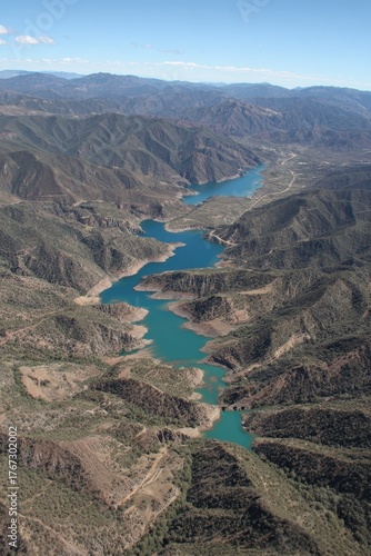Aerial view of a winding blue lake flowing through rugged arid tree covered mountains under a clear blue sky