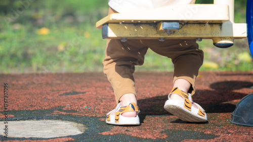 A cute rear view of a child's dangling feet in tiger-patterned sandals just above the red rubber playground floor. The image highlights freedom, fun, and movement in an outdoor park setting.