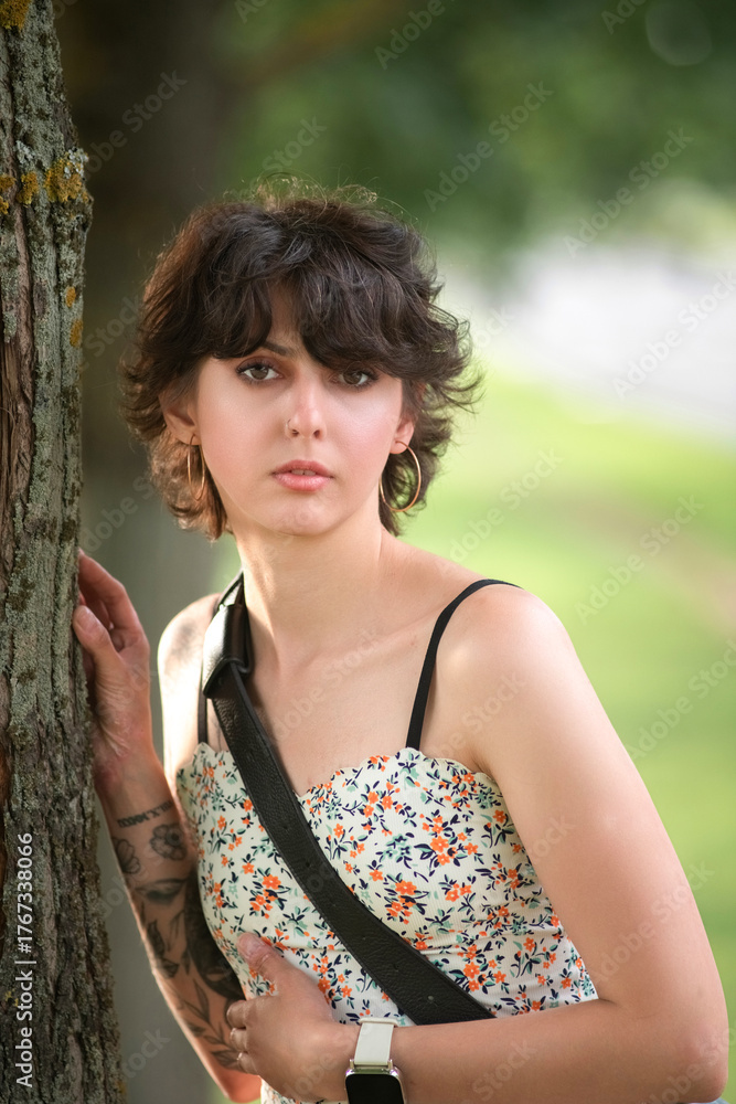Naklejka premium Young woman with short hair and floral dress leans against a tree, showcasing her tattoos and relaxed demeanor in a natural outdoor setting, capturing a moment of tranquility