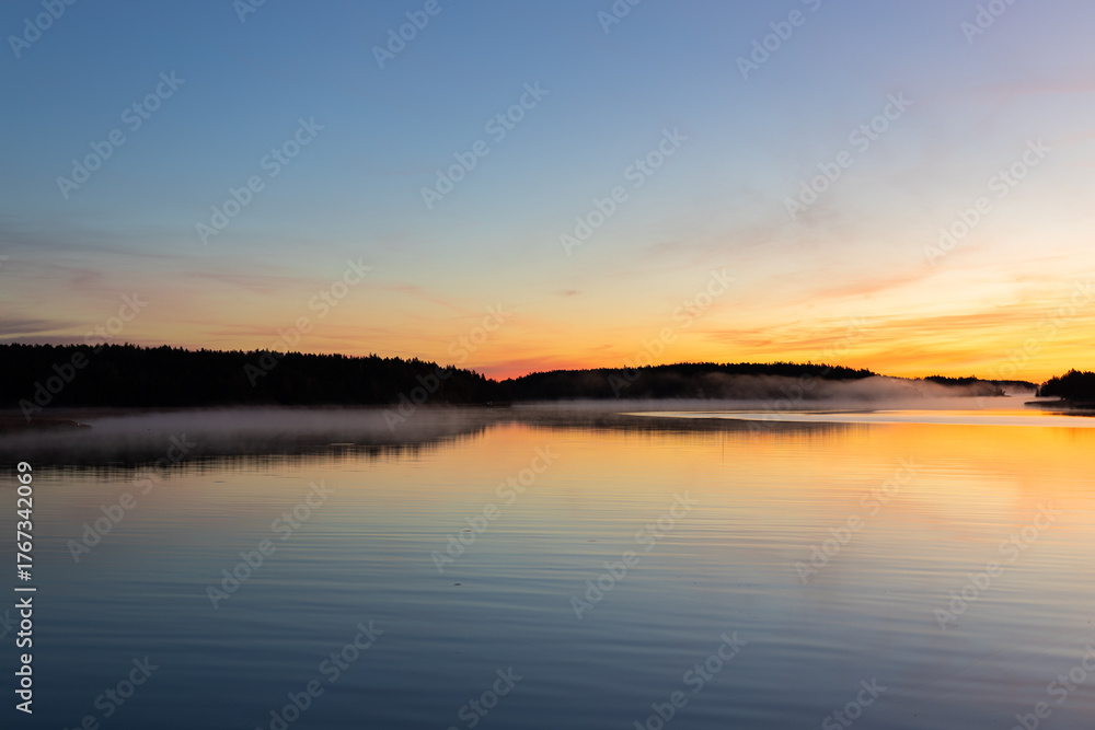 Obraz premium Golden sunrise over calm autumn sea with forest on the shore and gentle fog above the water, peaceful natural seascape and morning atmosphere