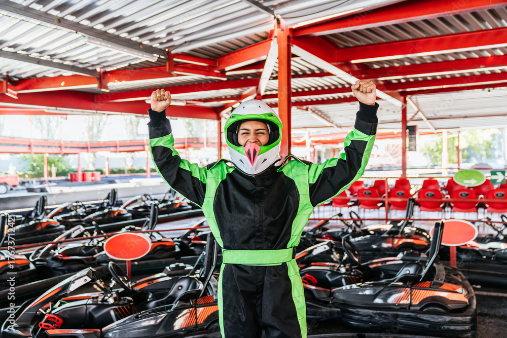 Obraz premium Young woman in a racing suit and helmet raising her arms, celebrating winning a go kart race at an outdoor track