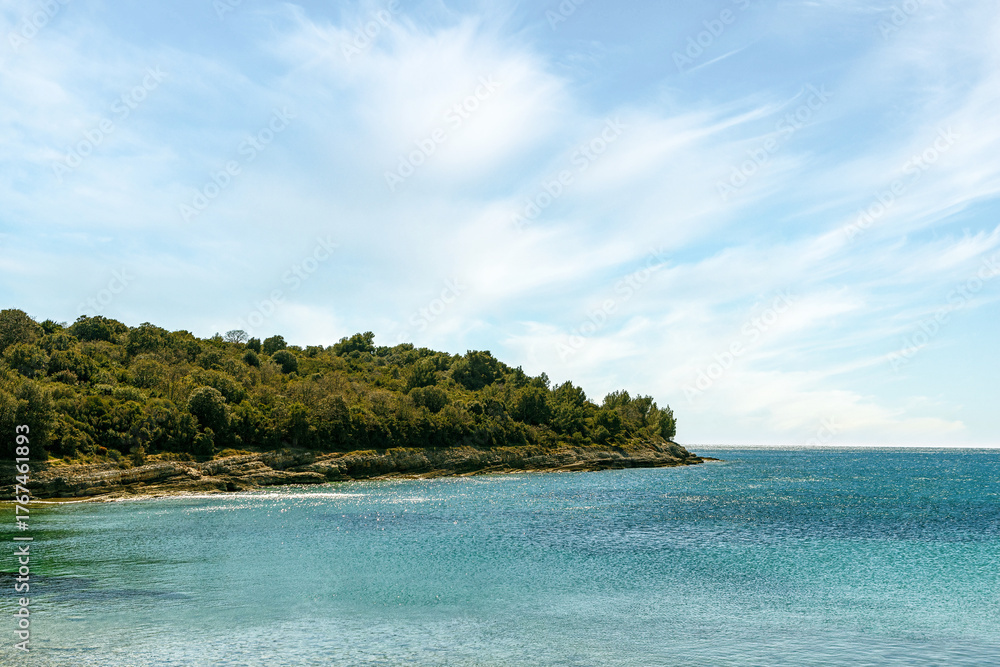 Naklejka premium View of a peaceful bay with anchored boats and rocky coastline in Cape Kamenjak Nature Park near Premantura, Croatia. The turquoise water contrasts with the stone shore and pine forest.
