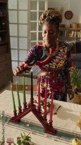 Vertical view of spiritual African American young woman wearing traditional dress lighting kinara while getting ready for Kwanzaa party