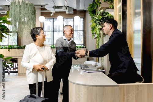 Happy senior Asian couple checking in at hotel on holiday vacation with young male receptionist. Receptionist man in suit shaking hands with Elderly traveler customer at hotel reception counter desk.
