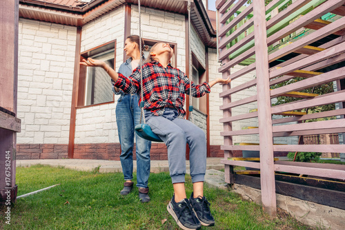 A young Caucasian boy swings joyfully in a backyard while a young Caucasian woman watches him. They share a moment of connection and happiness in a family setting.