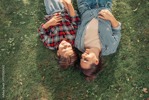 Two women lie on green grass, smiling and enjoying each other's company. One has long brown hair and wears a plaid shirt, the other has medium-length brown hair and a striped shirt.