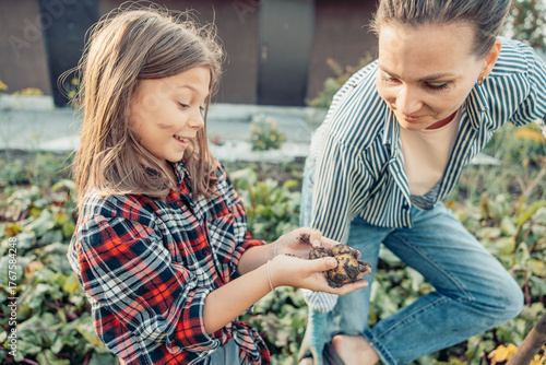 A young girl and a woman share a moment in a garden. The girl has long brown hair and wears a red plaid shirt. The woman has brown hair and wears a striped shirt. They are smiling and holding soil.