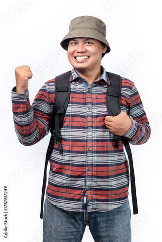 A man smiles wearing a travel look, with a bucket hat, flannel shirt, jeans, and black bag with a white background.