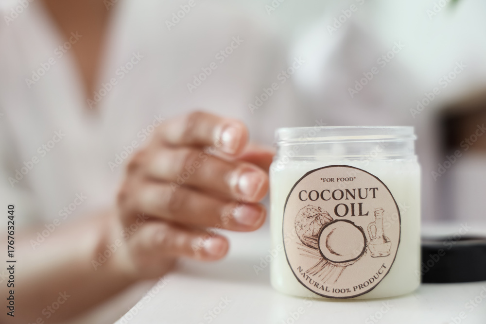 Fototapeta premium Young woman with jar of coconut oil on table in bedroom, closeup
