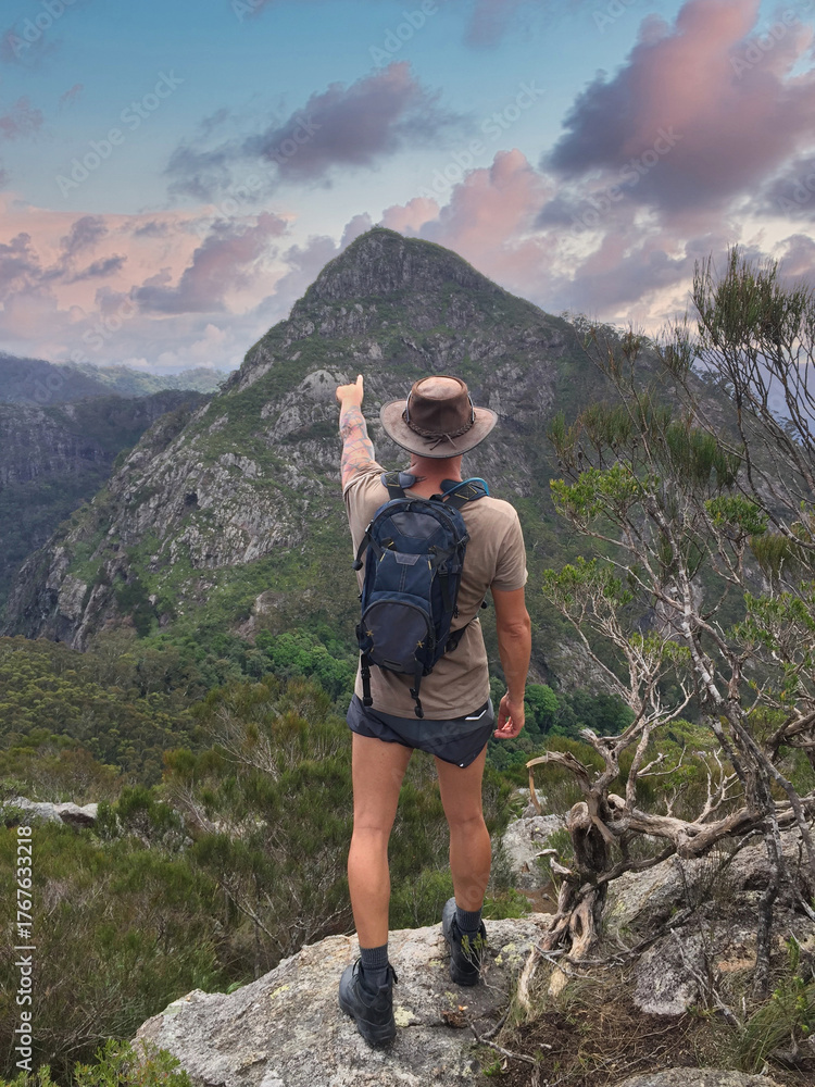 Obraz premium Back view of a lone Brazilian hiker with a hat and backpack standing on a rocky outcrop, pointing toward a distant mountain peak in the forested highlands of Australia under a colorful sky.