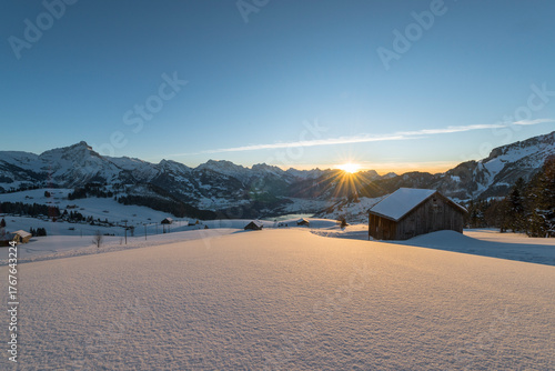 Verschneite Bergwiesen mit Heuhütten vor Panorama der Glarner Alpen bei Sonnenuntergang, Kanton Sankt Gallen, Schweiz