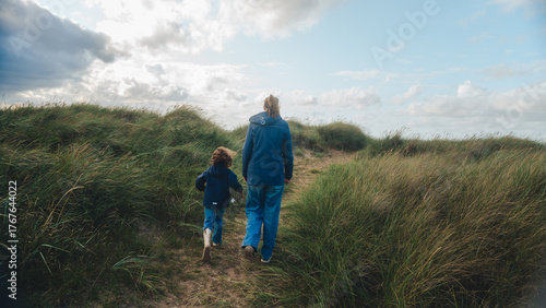 Fototapeta Naklejka Na Ścianę i Meble -  A mother and her child walk hand in hand through grassy sand dunes under a partly cloudy sky. The scene captures a peaceful moment of connection and exploration in nature, with soft natural light and 