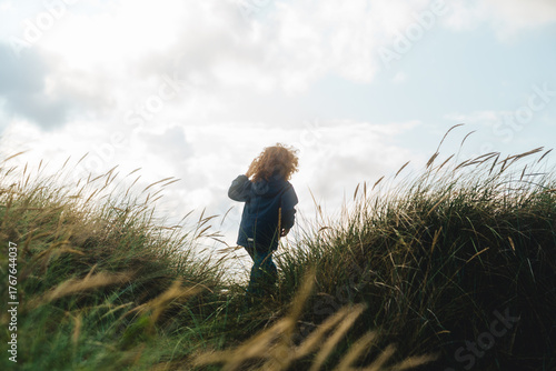 Fotografie A young child with curly hair walks through tall dune grass under a bright, cloudy sky