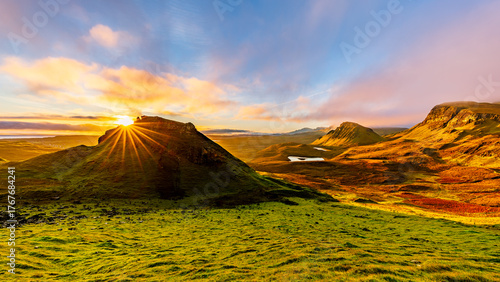 Breathtaking sunrise over Quiraing on the Isle of Skye, Scotland, where golden sunlight illuminates the dramatic green hills and valleys under colorful morning clouds.