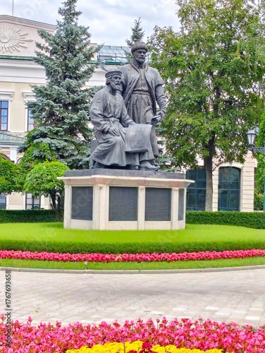   Kazan Kremlin,Kazan,Russia,July 26,2024:
 Kul Sharif.A bronze statue of two symbolic medieval builders with architectural plans,set in a green garden surrounded by flowers and historic buildings.