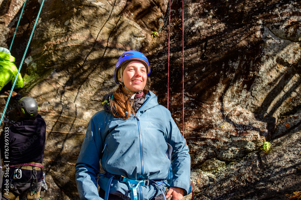 Naklejka premium Rock climber (young woman, in blue helmet, hardshell jacket, and blue climbing harness) standing in front of a rock face with belay line hanging vertically.