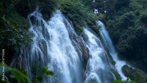 Lush Green Tropical Forest With Cascading Waterfall Flowing Over Mossy Rocks Amidst Dense Foliage In Soft Natural Light