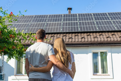Young couple embracing, looking at solar panels on the roof of their house