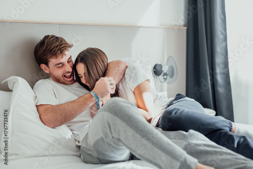 A young couple lies in bed, laughing and playfully teasing each other. Their smiles and playful touches show joy, closeness, and love, sharing a fun, relaxed moment at home.