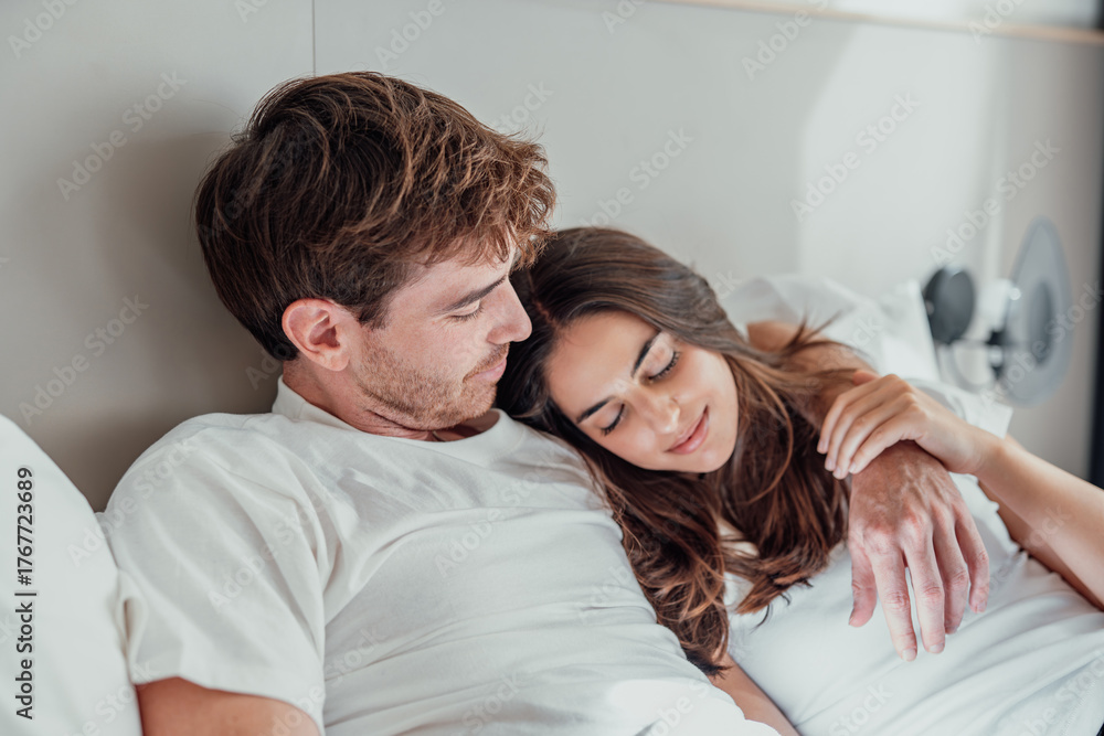 Fototapeta premium A young couple lies in bed, embracing and talking quietly. Their smiles and gentle touches show comfort, closeness, and love, sharing a peaceful moment in their cozy home.