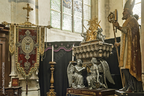 Quadro su tela large statue of a bishop and two kneeling angels a pedestal Behind them is a decorative banner with Latin text and a large cross inside St