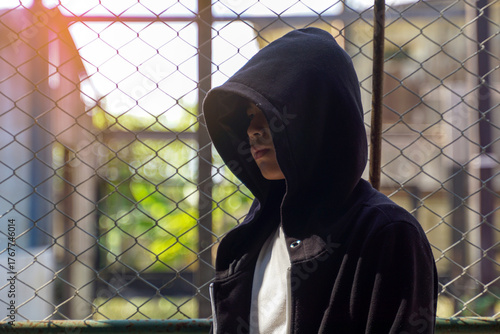 Mysterious teenage boy in a black hoodie standing in front of a chain-link fence. Urban youth, crime, and anonymity concept.