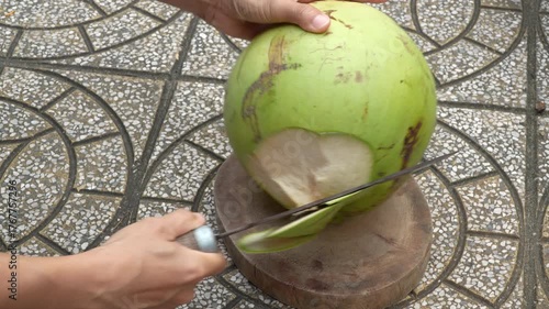 A man's hands chop a fresh green coconut with a large knife (machete) on a wooden block. Traditional way of opening a tropical coconut drink outdoors.