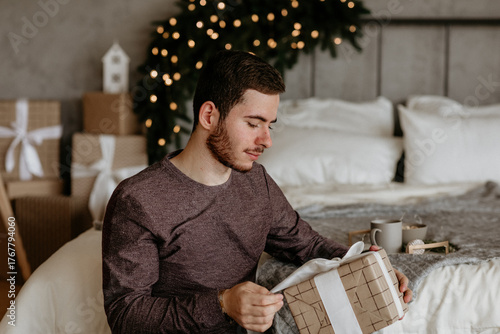 Young man in a casual sweater sitting in a cozy bedroom, carefully unwrapping a Christmas gift wrapped in brown paper and white ribbon, with blurred holiday decor in the background
