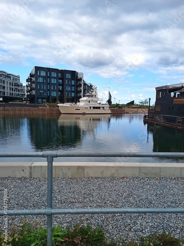 A modern yacht moored in a calm marina surrounded by contemporary residential buildings. Reflections on the water, partly cloudy sky, and peaceful urban harbor atmosphere. C