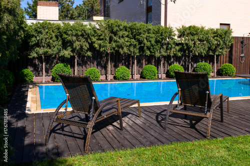 A spacious backyard with a swimming pool and two wicker loungers on a wooden deck. Surrounded by green shrubs, trees, and a modern privacy fence for a relaxing ambiance