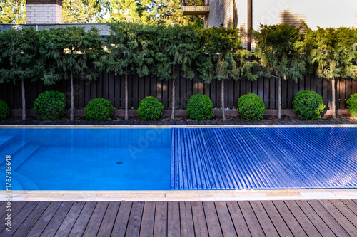 A partially covered outdoor swimming pool with a blue slatted pool cover in a private backyard. The area is surrounded by well-maintained shrubs, conifer trees, and a dark wooden fence