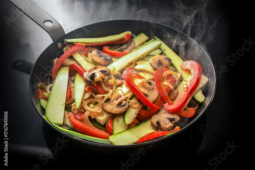 Stir fried vegetables in a steaming frying pan with red bell pepper, zucchini, mushrooms and onion on a black induction hob, vegetarian cooking concept, copy space, selected focus