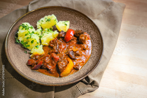 Hungarian style pork goulash with bell pepper served with potatoes and parsley garnish on a brown plate and a wooden table, savory meat dish, copy space, selected focus