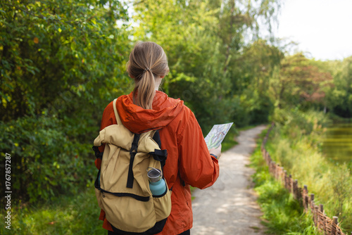 Rear view of young female traveler wearing orange jacket and carrying beige backpack while walking along forest trail with a paper map. Concept of adventure, exploration, hiking, navigation, and